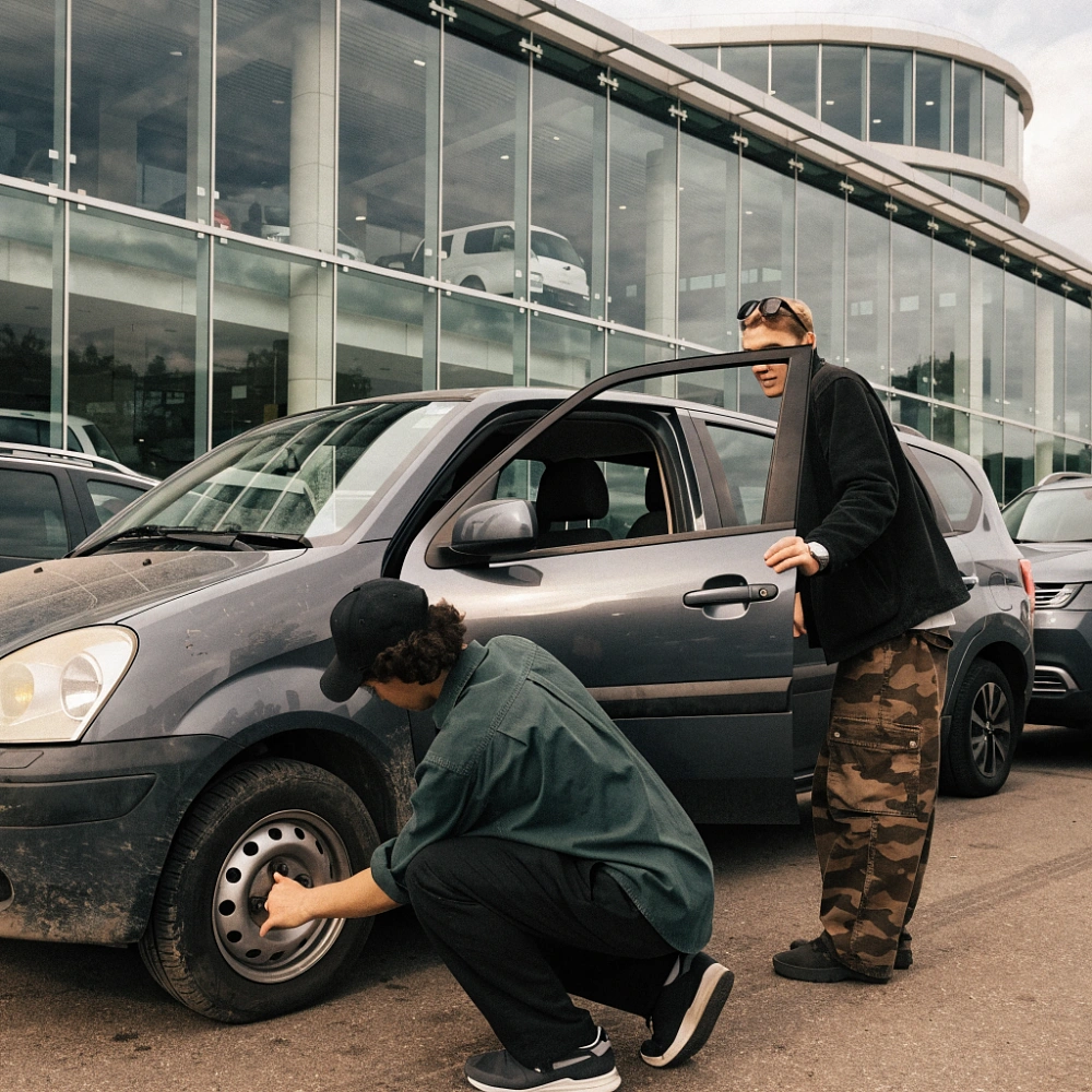 Двое осматривают подержанный компактный автомобиль на площадке у автосалона
